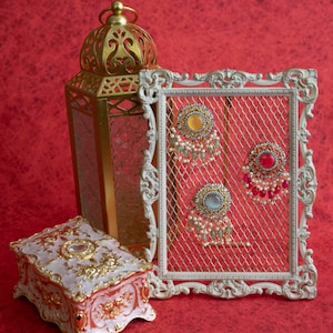 May include: Three pairs of gold earrings with colorful stones and pearl accents displayed on a white metal stand with a mesh background. The earrings are on a red surface with a gold lantern and a decorative box.