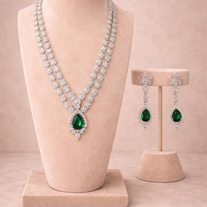 Pode incluir: Um deslumbrante conjunto de colar e brincos com cristais transparentes e pedras preciosas verdes esmeralda em forma de gota. O colar é exibido em um manequim e os brincos em um suporte. As joias estão em um fundo rosa suave.