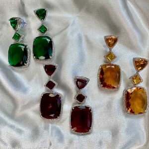 May include: Four pairs of dangle earrings featuring different coloured gemstones. Each earring has a square-shaped gemstone with a smaller triangular gemstone above it. The gemstones are green, orange, and red.