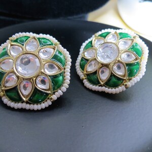 May include: A pair of round, green and gold earrings with white pearl accents. The earrings feature a floral design with a large, clear stone in the center.