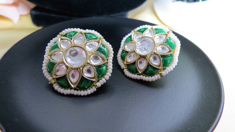 May include: A pair of round, green and gold earrings with white pearl accents. The earrings feature a floral design with a large, clear stone in the center.