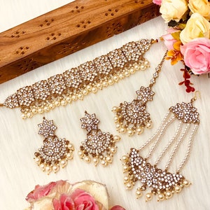 May include: A gold and white bridal jewelry set with a choker necklace, earrings, a maang tikka, and a long necklace with pearl accents.