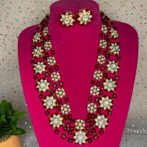 May include: A four-strand necklace with red beads and gold accents. Each strand features large, round, white stones with a floral design. The necklace is paired with matching earrings with red stones and gold accents.