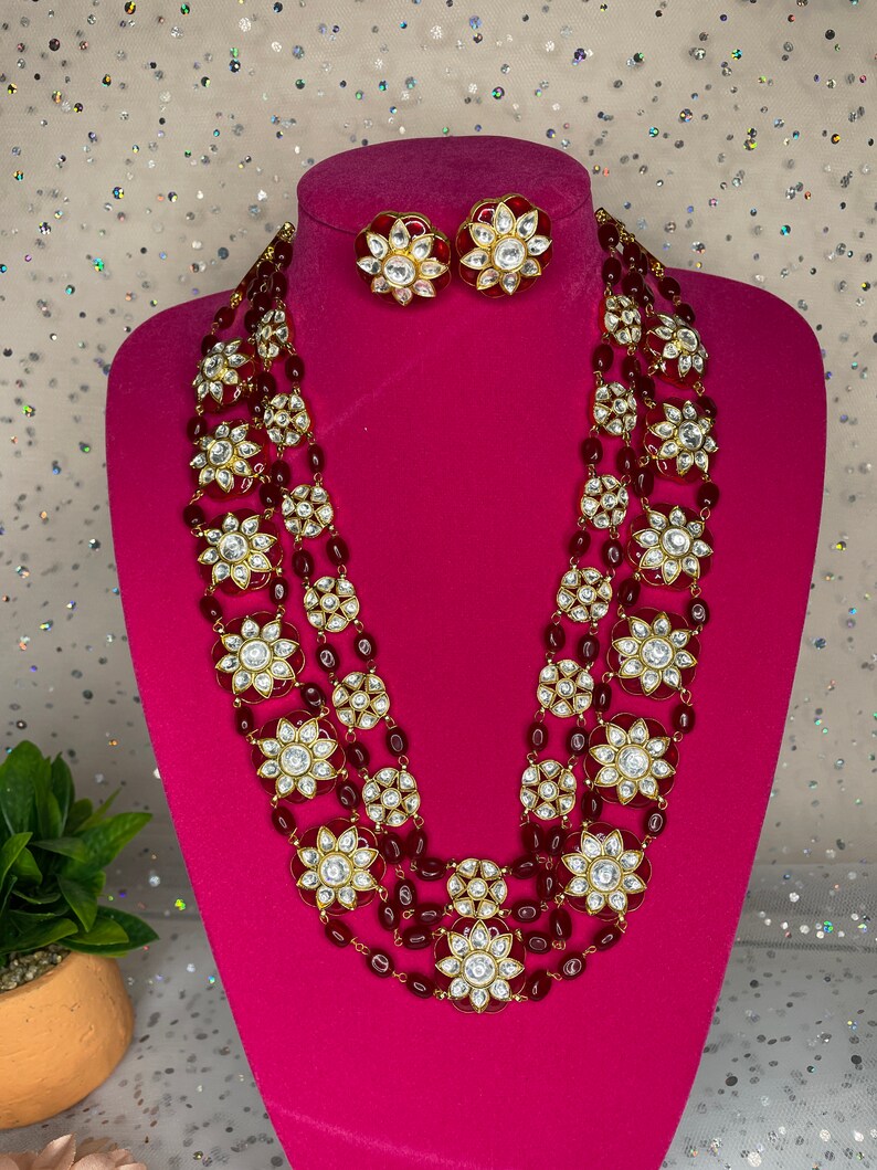 May include: A four-strand necklace with red beads and gold accents. Each strand features large, round, white stones with a floral design. The necklace is paired with matching earrings with red stones and gold accents.