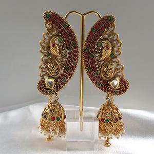May include: Ornate gold-toned earrings with a peacock design. The earrings feature red and green gemstones, a teardrop-shaped clear stone, and dangling bell-shaped pendants with faux pearls. The earrings are displayed on a gold-colored stand against a white background.