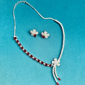 Può includere: Una collana d'argento con un ciondolo di pietra rossa e orecchini abbinati. Il ciondolo è un motivo a fiore con una catena pendente.
