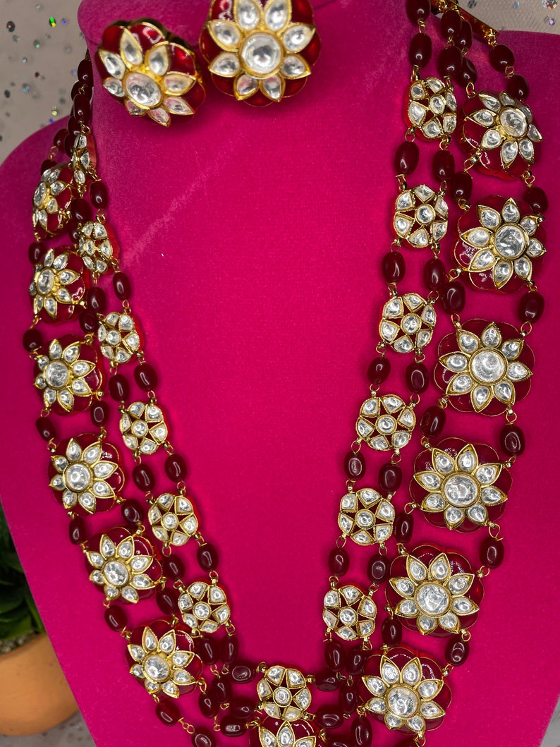 May include: A multi-strand necklace with red beads and gold-toned flower-shaped pendants with white stones. The necklace is paired with matching earrings.