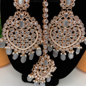 May include: A set of gold-tone earrings and a matching maang tikka, featuring clear and white stones. The earrings have a teardrop shape with dangling white beads. The maang tikka has a similar design with a central stone and dangling beads.