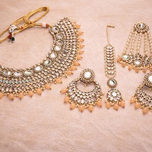 May include: A gold-toned bridal jewelry set featuring a necklace, earrings, a headpiece, and a tikka. The set includes reflective mirror accents and peach-colored bead drops. The pieces are arranged on a light peach surface.