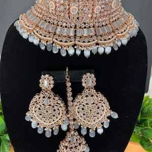 May include: A white and gold bridal jewelry set with a choker necklace, earrings, and a maang tikka. The set features white stones and pearls.