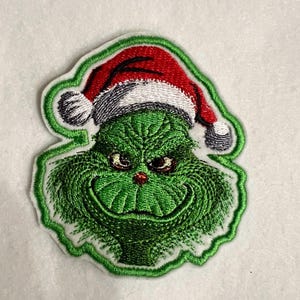 May include: Green Grinch character wearing a red and white Santa hat. The Grinch is smiling with a mischievous expression. The character is embroidered on a white fabric.