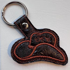 May include: A brown leather keychain with a red embroidered cowboy hat design. The keychain has a silver metal ring.