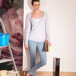 May include: A woman wearing a light purple long-sleeved shirt and blue jeans stands next to a wall with floral wallpaper. She is holding a brush and a roll of wallpaper.