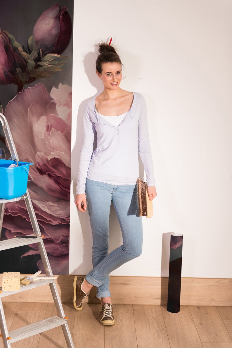 May include: A woman wearing a light purple long-sleeved shirt and blue jeans stands next to a wall with floral wallpaper. She is holding a brush and a roll of wallpaper.