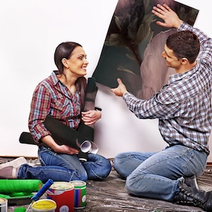 May include: A couple working on a home improvement project. The woman is holding a roll of wallpaper and the man is holding a large piece of artwork. They are both smiling and looking at each other.