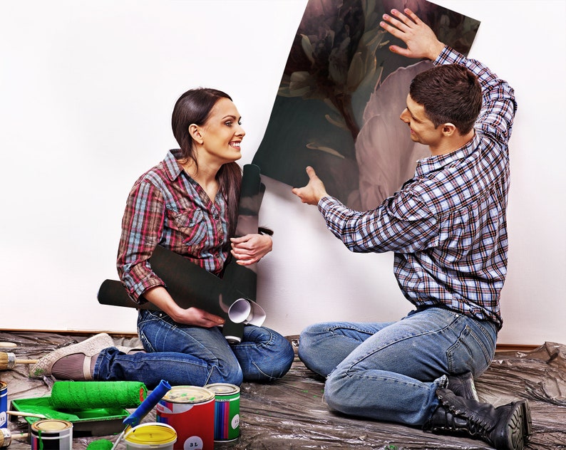May include: A couple working on a home improvement project. The woman is holding a roll of wallpaper and the man is holding a large piece of artwork. They are both smiling and looking at each other.