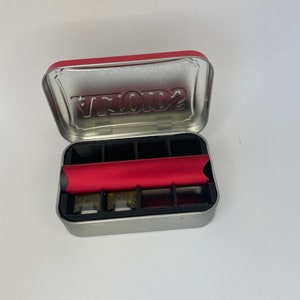 May include: A silver metal tin with a red lid. The lid has the word "VAPOR2" embossed on it. The tin is open and contains a red vape pen and several empty compartments.