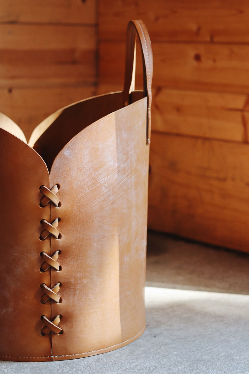 May include: A brown leather storage basket with a handle and a decorative stitched design. The basket is made of natural leather and has a rustic look.