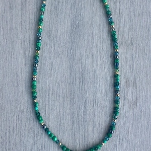 May include: A beaded necklace featuring a variety of green malachite-style beads, dark faceted crystals, and small gold-toned metallic spacers. The necklace is finished with a gold-toned clasp and a single green and gold-toned drop pendant.