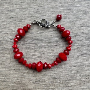 May include: A red beaded bracelet with a silver clasp. The bracelet features various sizes of faceted red beads. The clasp is a silver toggle style. The beads are arranged in a circular pattern.