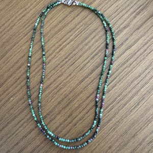 May include: A double-strand necklace featuring small, faceted beads in shades of green, black, and red. The necklace is secured with a silver-colored clasp. The beads are strung together to create a delicate and elegant piece of jewelry.