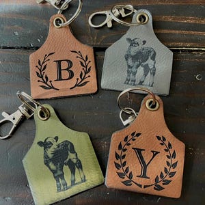 Peut inclure: Quatre porte-clés en cuir avec des fermoirs en métal. Deux porte-clés sont marron, un est gris et un est vert olive. Deux porte-clés ont une lettre noire dans un motif de couronne de laurier. Les deux autres ont une illustration de veau noir.