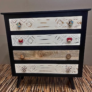 Op de afbeelding: Een dressoir met vier lades, een zwart frame en witte lades. Elke lade heeft een uniek gesneden ontwerp en kleurrijke knoppen. Het dressoir staat op een houten vloer.