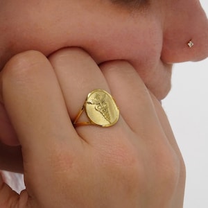 May include: A gold-colored ring with an oval face featuring a medical symbol. The ring is worn on a finger. The ring's design includes a staff with two snakes and wings. The ring is a piece of jewelry.