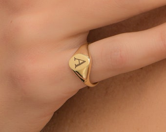 May include: A gold signet ring with the letter "A" engraved on the oval face. The ring is a warm, shiny gold color and is worn on a finger. The ring has a classic, minimalist design.