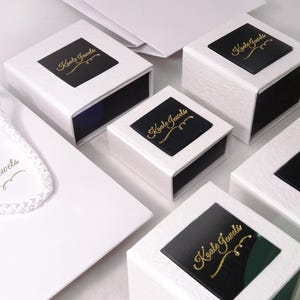 May include: Five white gift boxes with black lids. Each lid has a gold logo that says "Koala Jewels". The boxes are arranged in a staggered pattern on a white surface.