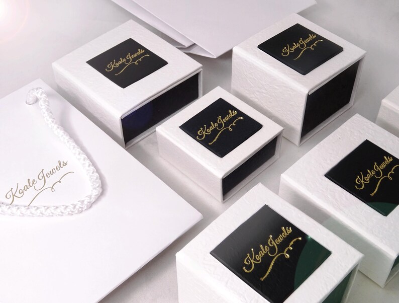 May include: Five white gift boxes with black lids. Each lid has a gold logo that says "Koala Jewels". The boxes are arranged in a staggered pattern on a white surface.