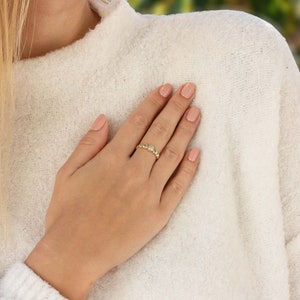 Peut inclure: Une bague en or avec une petite pierre précieuse transparente est portée au doigt. La bague présente un design délicat avec de petits ornements ronds. La personne porte un pull crème.