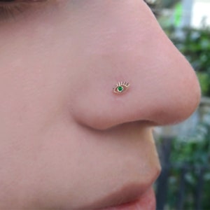 Könnte beinhalten: Ein goldener Nasenring mit einem grünen Edelstein in Form eines Auges mit Wimpern.