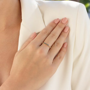 May include: A gold band ring with a row of colorful gemstones. The ring is on a person's finger.
