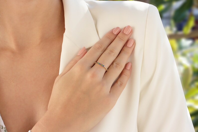 May include: A gold band ring with a row of colorful gemstones. The ring is on a person's finger.
