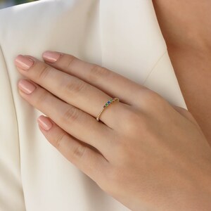 May include: A delicate gold band ring with a row of colorful gemstones. The ring is set on a finger with light pink nail polish.