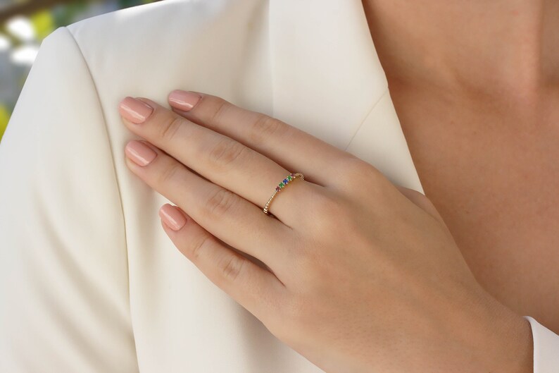 May include: A delicate gold band ring with a row of colorful gemstones. The ring is set on a finger with light pink nail polish.