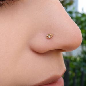 May include: A gold nose stud shaped like an eye with a central clear gemstone. The design features a gold outline of an eye with radiating lines representing eyelashes, centered with a small, round, clear stone as the pupil of the eye.