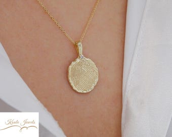May include: A gold-colored pendant necklace featuring a fingerprint design. The oval-shaped pendant has a textured surface and a small diamond accent. The necklace is worn against a white shirt.
