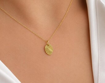 May include: A gold-toned necklace featuring a delicate chain and a small, oval-shaped pendant. The surface of the pendant is engraved with a detailed, circular fingerprint pattern that radiates from the center, creating a textured appearance.