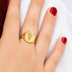 May include: A gold-toned ring featuring a flat, oval-shaped face with an engraved caduceus symbol. The design displays a central staff entwined by a single snake with wings at the top. The band is thin and splits into two sections near the face.