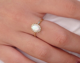 Könnte beinhalten: Ein goldener Ring mit einem ovalen Opal-Edelstein, umgeben von kleinen, klaren Akzentsteinen. Der Ring ist auf einem zarten Goldband gefasst. Der Opal hat ein milchig-weißes Aussehen mit subtilen irisierenden Blitzen.