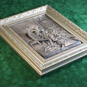 May include: A silver framed wall art piece depicting a three-dimensional sculpture of a woman holding a crescent moon.