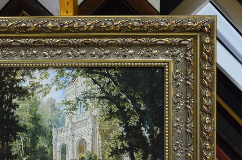 May include: A gold-toned ornate picture frame with a detailed design. The frame surrounds a painting of a white building with trees and a path leading to the building.