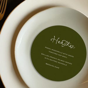 May include: A formal place setting with a gold fork and white plates with gold rims. A green circular menu card with the name "Heather" and a list of menu items is centered on the plate.