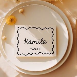 May include: A white place card with a black wavy border and the text "Kamila Table 2" on a white plate with gold silverware and a lemon on a beige tablecloth.