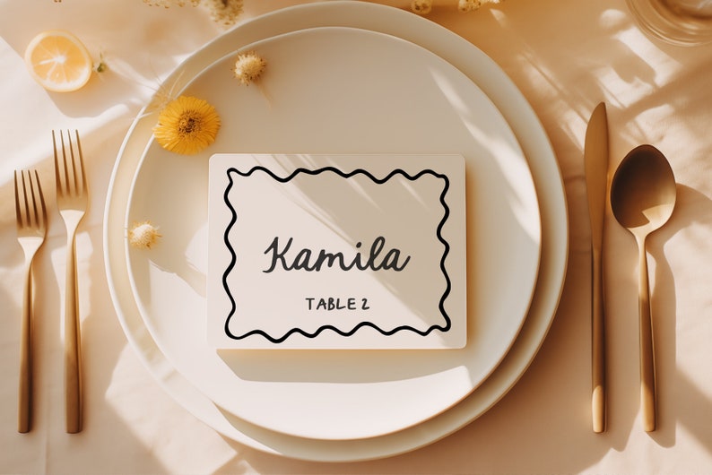 May include: A white place card with a black wavy border and the text "Kamila Table 2" on a white plate with gold silverware and a lemon on a beige tablecloth.
