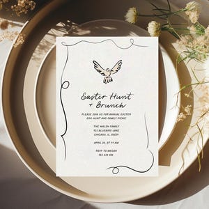 May include: A white card with black calligraphy outlining a dove with a pink body. The text reads "Easter Hunt + Brunch" and includes details for an annual Easter egg hunt and family picnic. The card is on a beige plate with a white background.