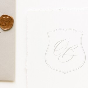 Puede incluir: Una tarjeta blanca con una forma de escudo gris y las iniciales "OB" escritas en cursiva dentro del escudo.