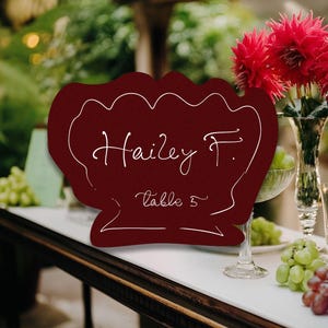 May include: A burgundy-colored place card with the name "Hailey F." and "table 5" in white script. The card has a decorative scalloped edge. The background includes red flowers, grapes, and glassware, suggesting a wedding or event setting.